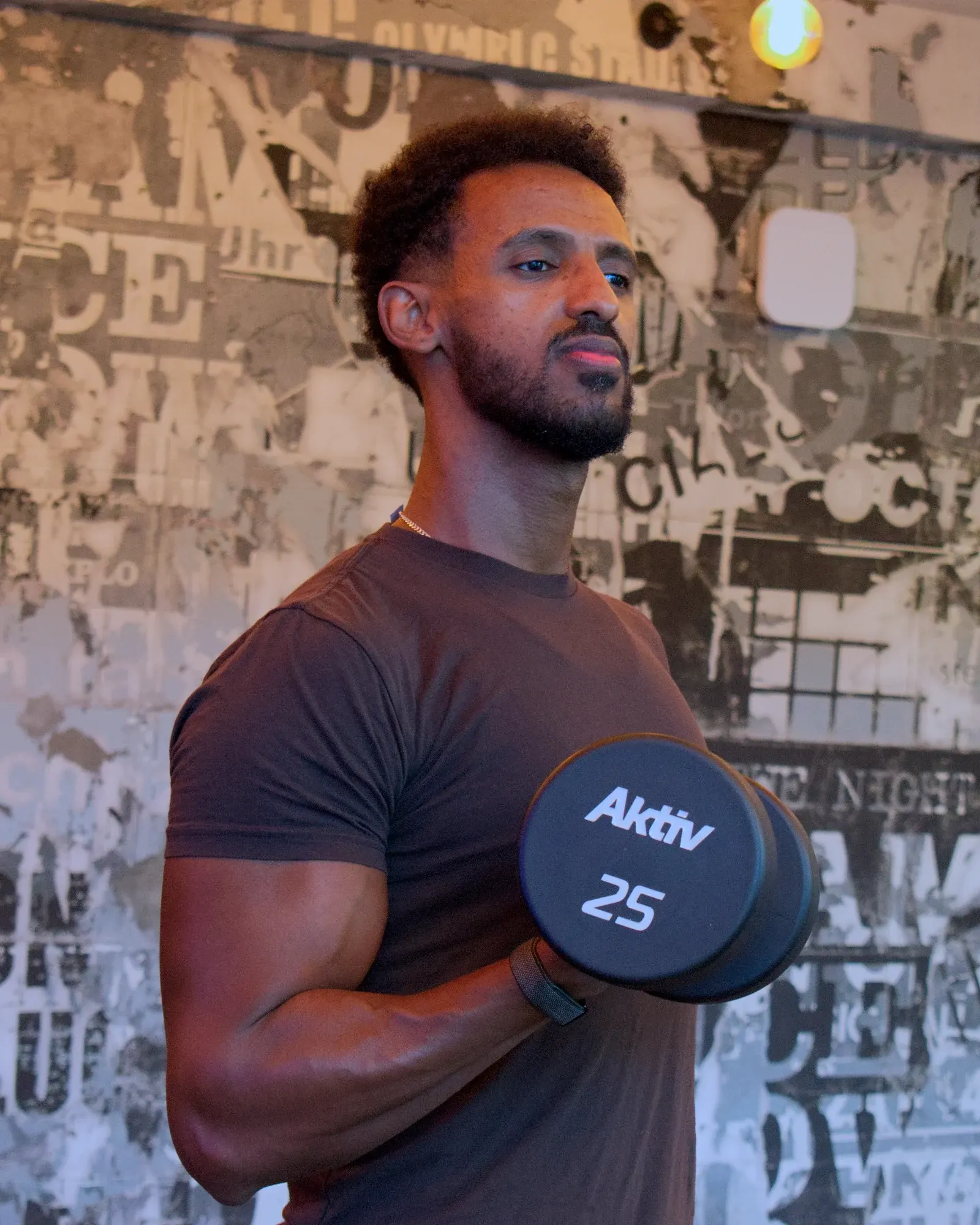 Nathnael Gebre performing a dumbbell biceps curl in front of a black-and-white poster wall.