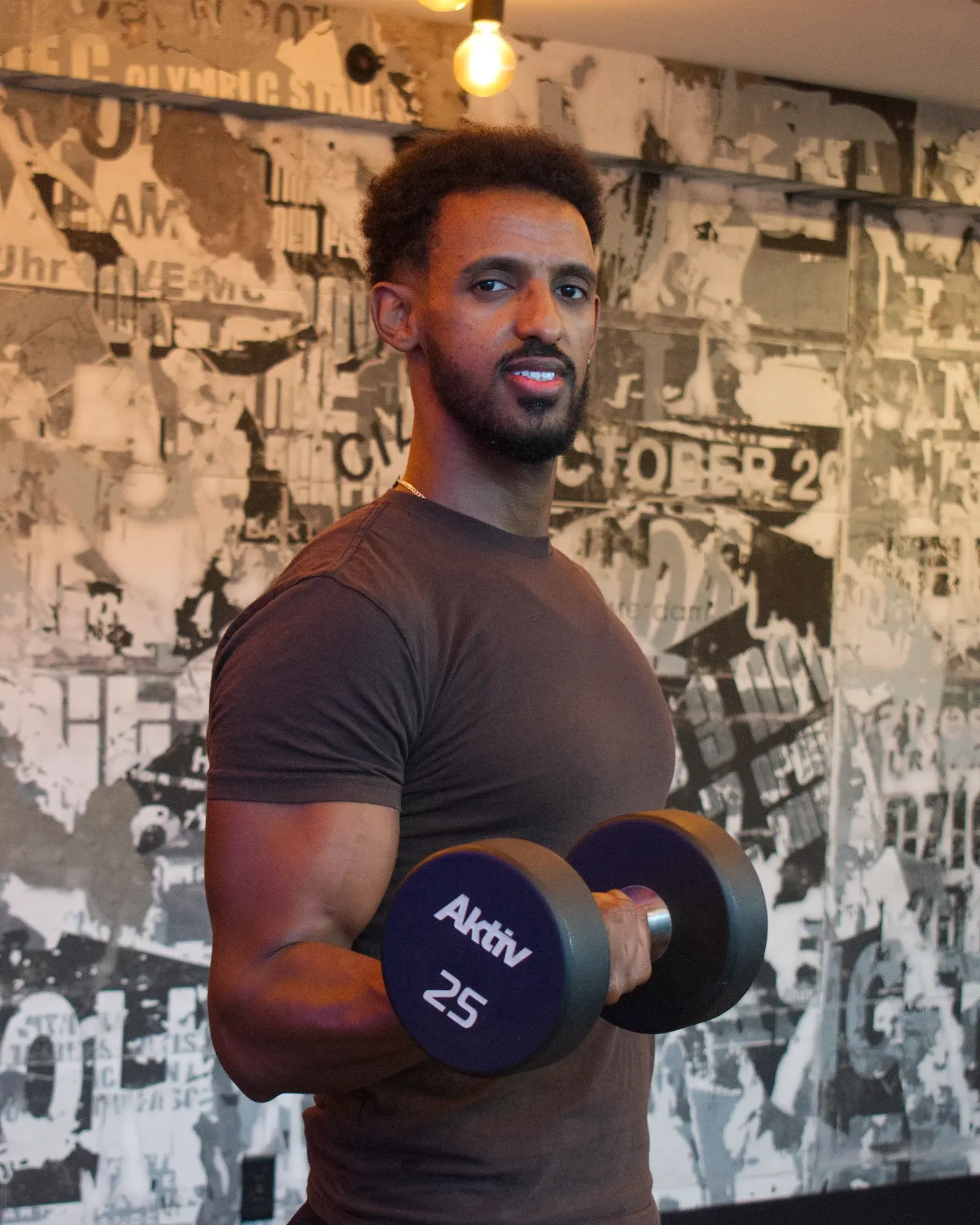 Nathnael Gebre smiling at the camera during a dumbbell curl in front of a poster-covered gym wall.