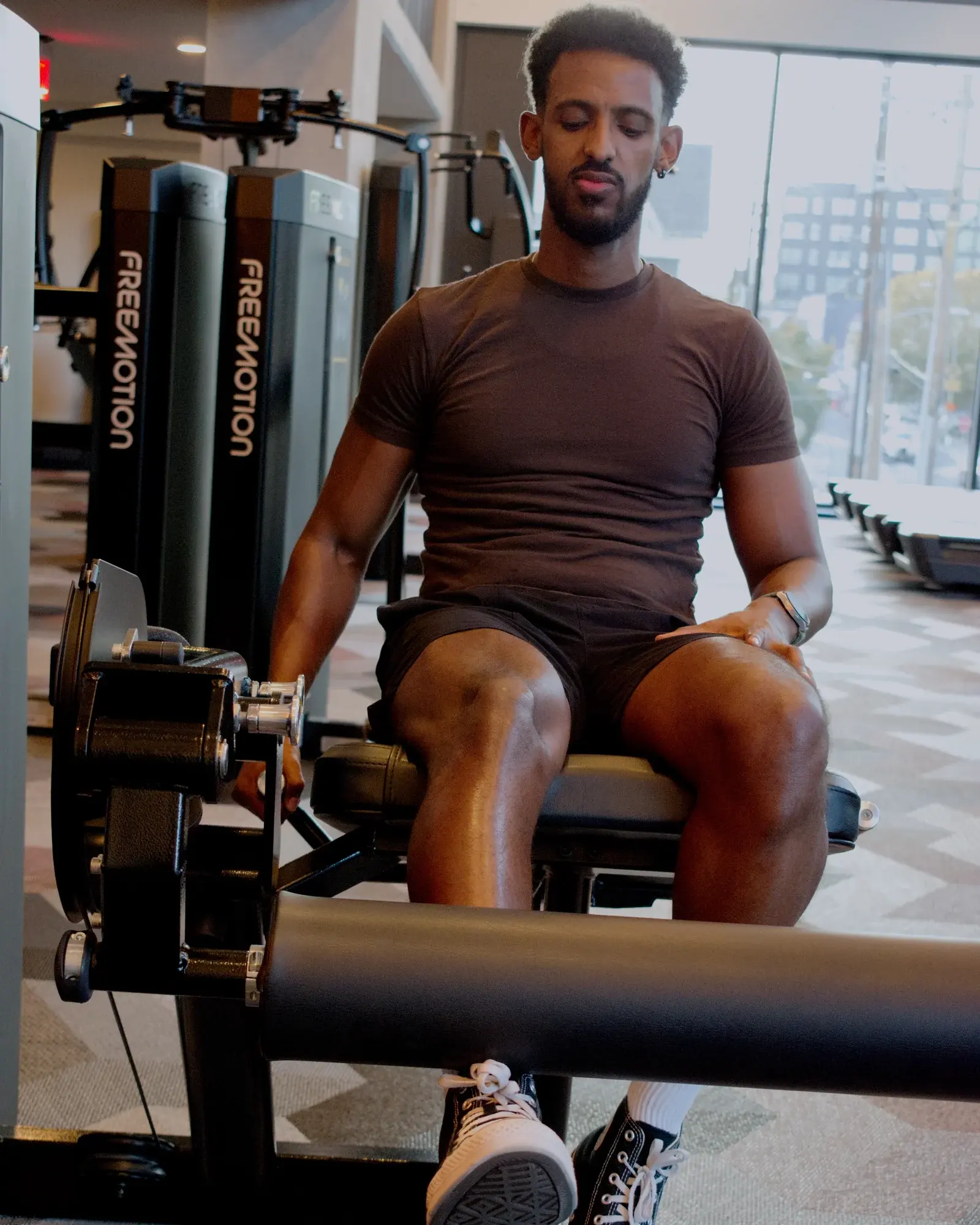 Nathnael Gebre seated on a leg extension machine in a bright gym with floor-to-ceiling windows.